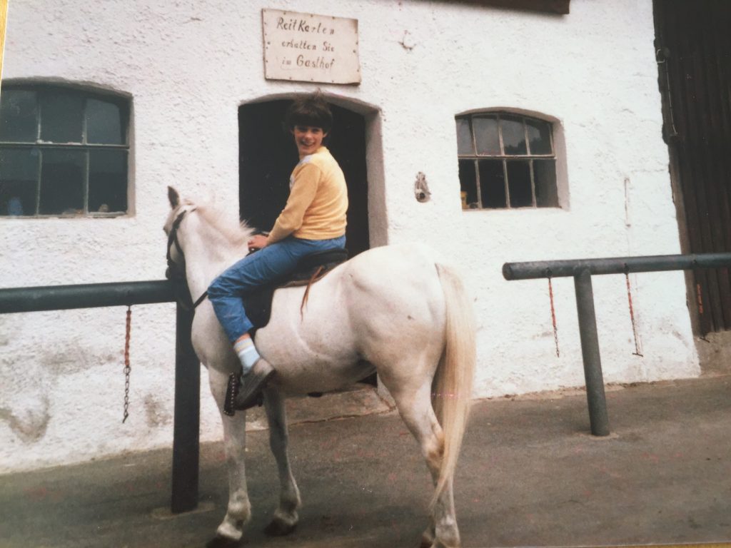 Carola sitzt auf einem weissen Pferd. Sie steht mit dem Pferd vor dem Stall.