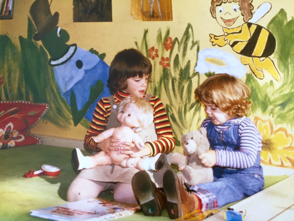 Carola und ihre Schwester Sonja als kleine Mädchen. Sie sitzen auf dem Boden und spielen mit einer Puppe und einem Teddynbären.