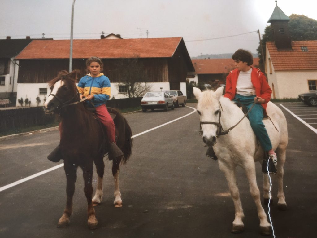 Carola und ihre Schwester Sonja sitzen auf Pferden. Sie reiten auf einer Strasse.