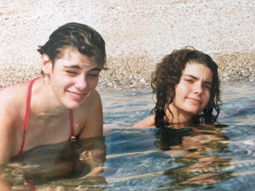 Carola und ihre Schwester Sonja am Meer. Sie sitzen beide im Wasser. Nur noch der Kopf ist zu sehen. Sie lächeln.