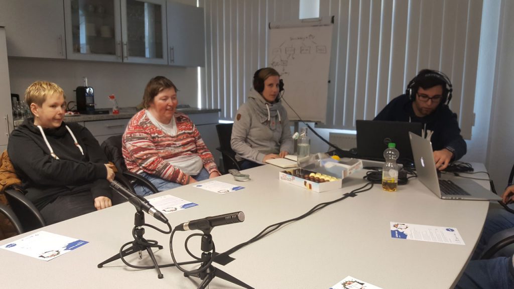 Die Teilnehmer am Podcast sitzen vor Mikrofonen, die auf dem Tisch positioniert sind.