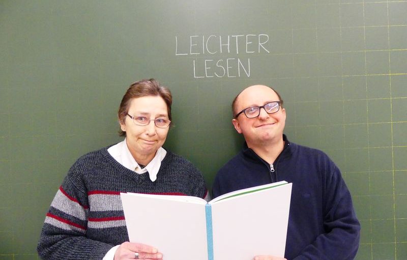 Monika Birk und Robert Gubitz stehen lächelnd vor einer grünen Schultafel. Darauf steht: Leichter lesen. Die beiden halten ein aufgeschlagenes Buch.