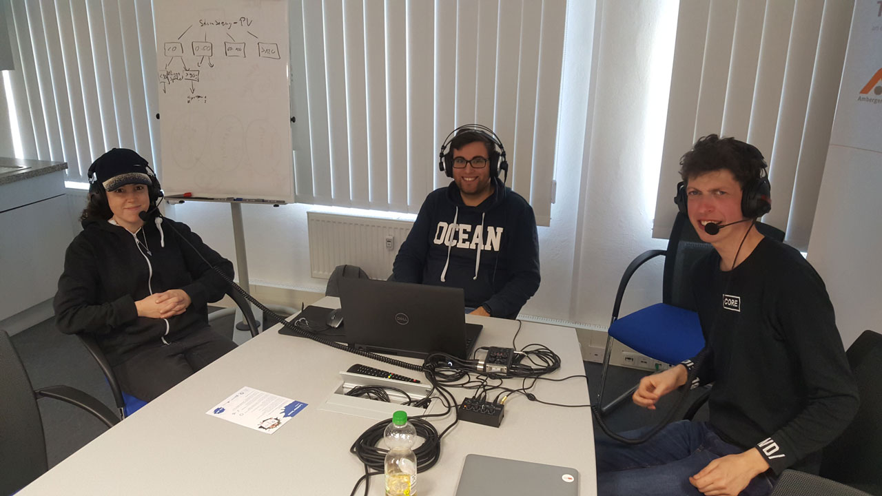 Auf dem Foto sitzt Janine links, Philipp rechts und in der Mitte Simon. Alle drei tragen Kopfhörer und bereiten sich auf eine Podcast-Aufnahme vor.