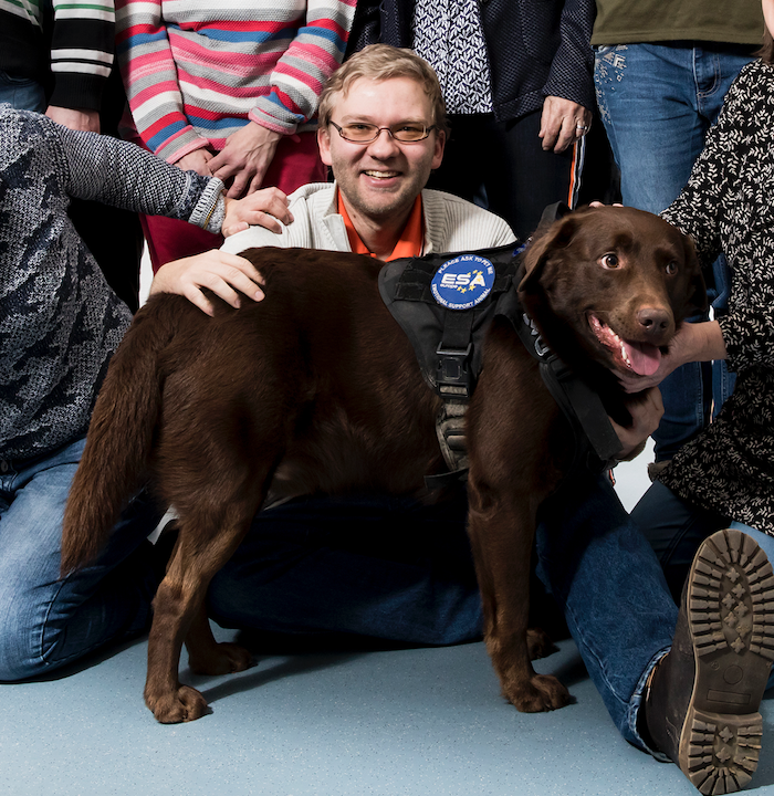 Andreas sitzt auf dem Boden und lächelt über den Rücken seines Hundes Janosch in die Kamera.