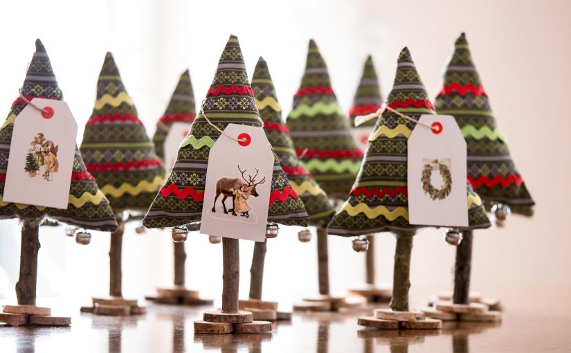 Viele bunte Christbaume aus Stoff mit Verzierungen auf einem kleinen Astständer. An den Bäumen hängen kleine Geschenkkärtchen.